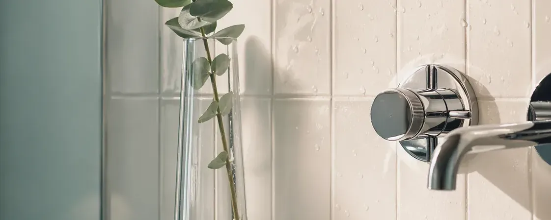 A pristine white subway-tiled bathroom corner with polished chrome fixtures, water beading on tile, and a single eucalyptus stem in a clear vase
